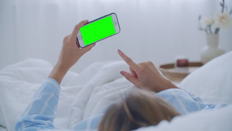 Young-woman-with-cell-phone-on-bed-shoulder-view.-Technology-addiction.-Green-screen-phone.-Young-woman-touching-belly-and-using-smartphone-in-bedroom